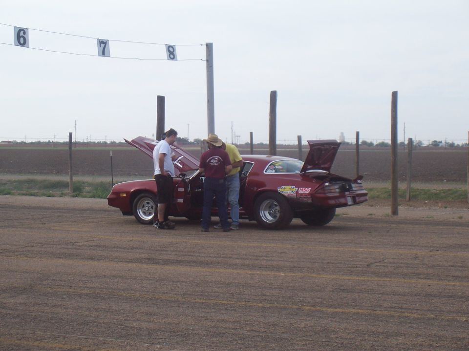 Red coupe with hood and trunk open, three people inspecting the engine bay, drag racing stickers on the rear fender