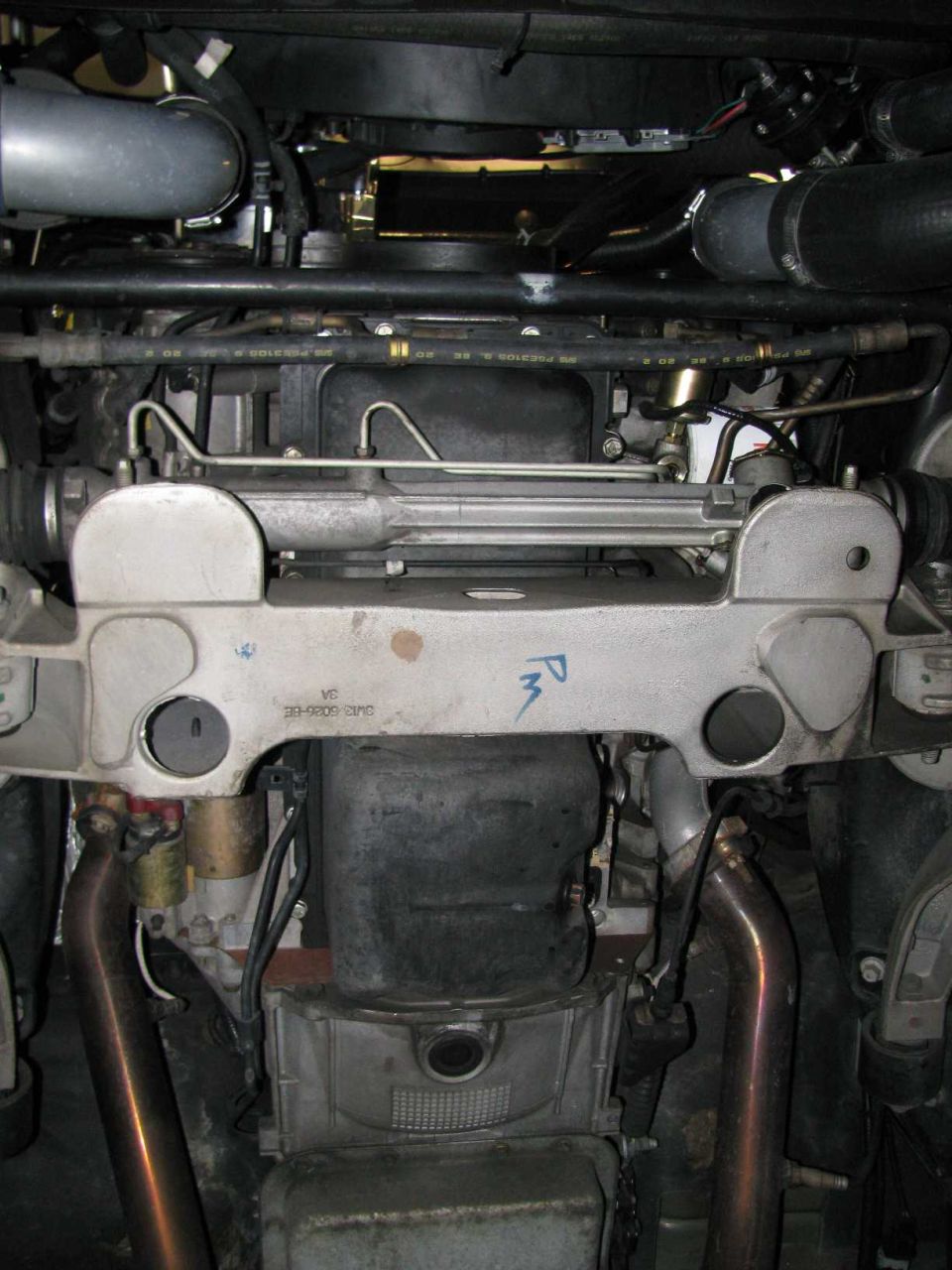 Underbody view of a V8 engine showing aluminum crossmember, steering rack, oil pan, starter, and dual exhaust pipes