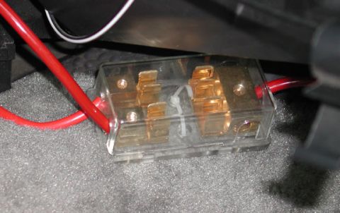 1988 Ford LTD Crown Victoria - Close-up of a clear-housing gold-plated ANL/AGU distribution fuse block mounted to gray carpet under a vehicle's interior trim, with heavy-…