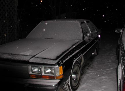 1988 Ford LTD Crown Victoria - Nighttime three-quarter front view of a dark boxy 1980s Ford full-size sedan parked in falling snow, its hood, roof and trunk covered in a…