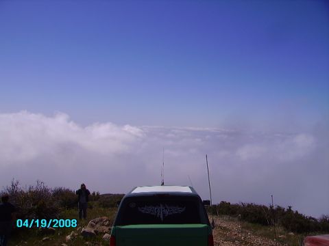 1990 Ford Bronco - A green SUV is parked on a hilltop overlooking a sea of clouds, with two people visible in the distance.