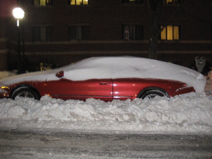 Red car buried in snow, side view. Snow covers most of the vehicle, obscuring details