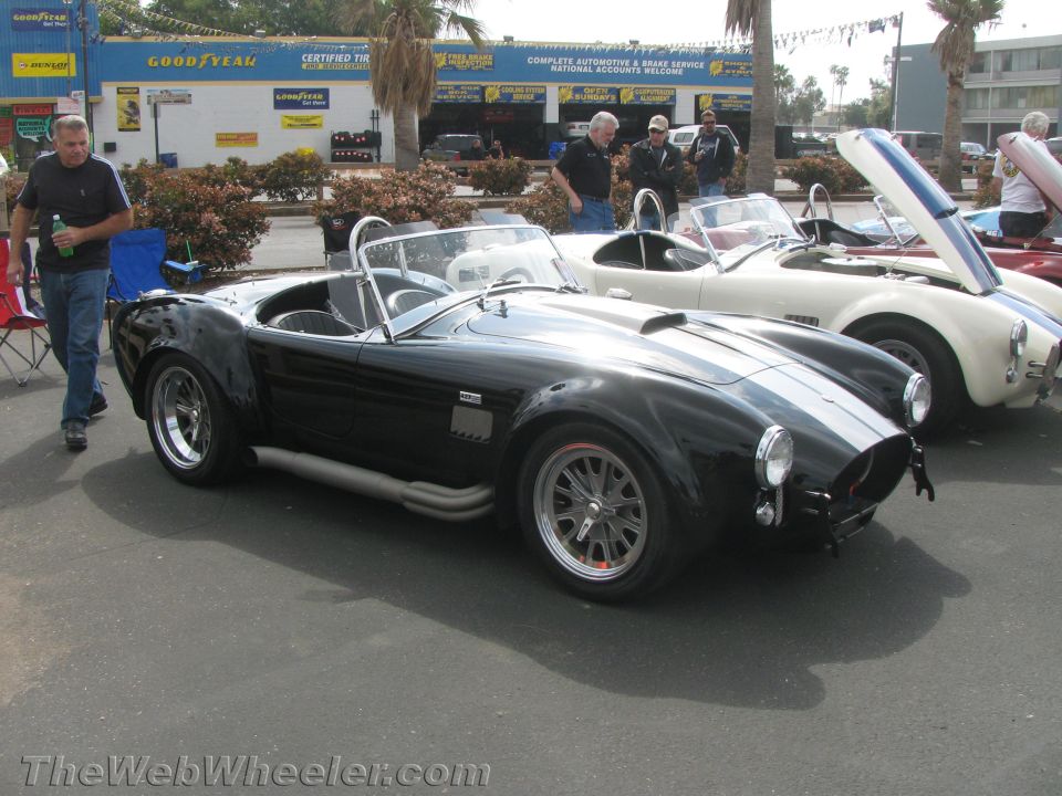 Black AC Cobra sports car with dual white racing stripes and side exhaust pipes. White Cobra behind it
