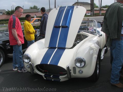 1968 Ford Mustang - A white Shelby Cobra replica with blue racing stripes is displayed with its hood open, attracting attention from onlookers.