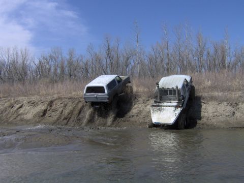 1992 Ford F-350 - Two off-road vehicles navigate a muddy water crossing with a dirt embankment on the side.