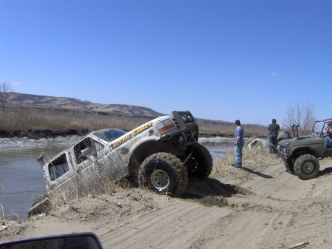 1992 Ford F-350 - Two off-road trucks are stuck in the sand near a body of water under a clear blue sky.