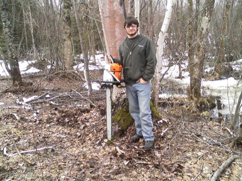 2000 Chevrolet Tahoe - A man stands in a wooded area with a chainsaw, ready for work. The ground is covered in leaves and patches of snow.