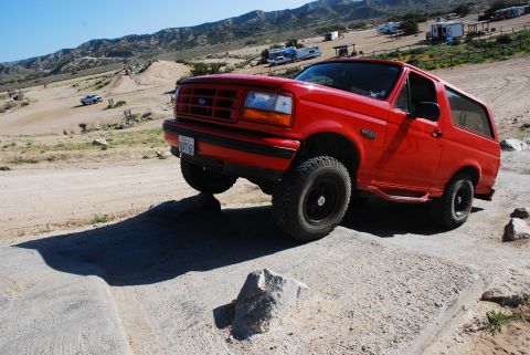 1995 Ford Bronco - A red Ford Bronco navigates an off-road obstacle course with other vehicles and RVs in the background.