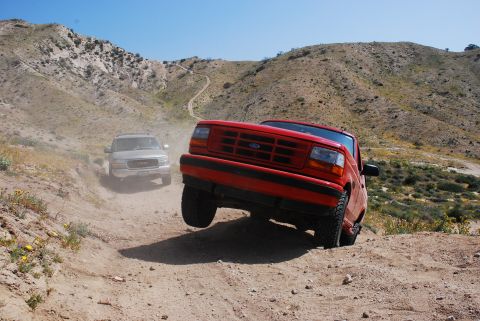 1995 Ford Bronco - A red 1995 Ford Bronco navigates a dusty trail in a hilly, arid landscape, with another SUV following behind.