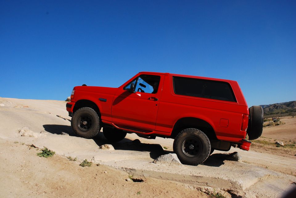 Red 1995 Ford Bronco climbing over rocks on a dirt trail under a clear blue sky