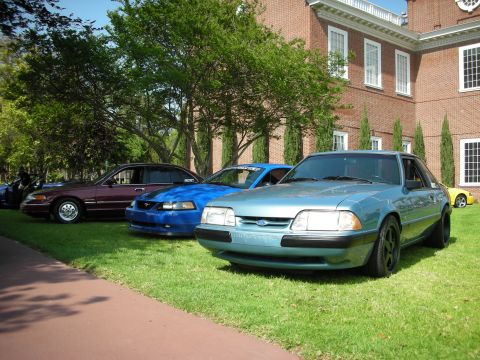 1992 Ford Crown Victoria - Three Ford Mustangs are parked on a grassy lawn in front of a large brick building.