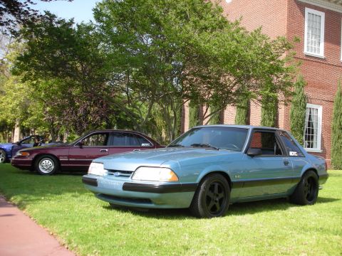 1992 Ford Crown Victoria - A blue coupe with black wheels is parked on the grass in front of a brick building, with other vehicles visible behind it.