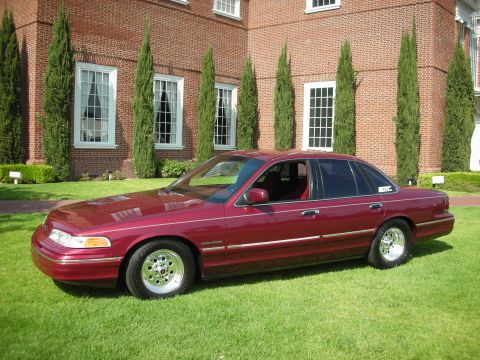 1992 Ford Crown Victoria - A maroon 1992 Ford Crown Victoria sedan is parked on a lawn in front of a large brick building with tall windows.