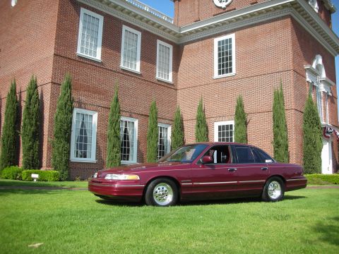 1992 Ford Crown Victoria - A maroon sedan is parked on a grassy lawn in front of a large brick building with white-trimmed windows.