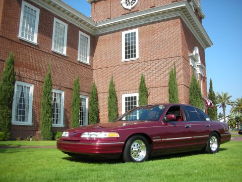 1992 Ford Crown Victoria - A maroon 1992 Ford Crown Victoria is parked on a lawn in front of a large brick building with white-trimmed windows.