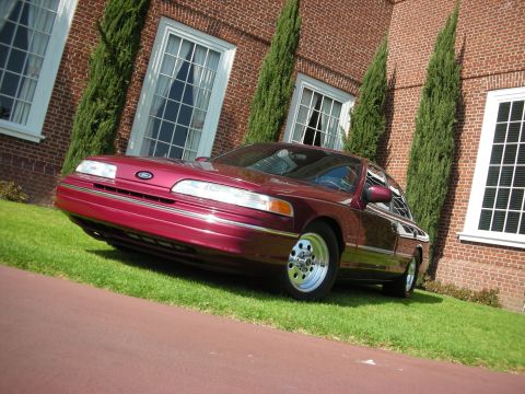 1992 Ford Crown Victoria - A red 1992 Ford Crown Victoria is seen from a 3/4 front angle, parked on a grassy lawn with a brick building in the background.