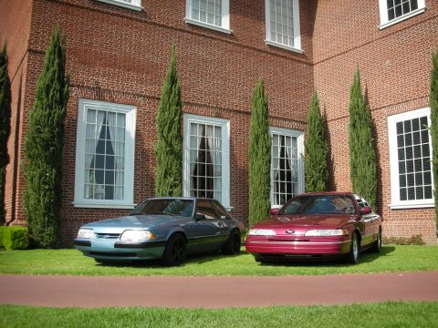 1992 Ford Crown Victoria - A maroon 1992 Ford Crown Victoria is parked on the lawn in front of a brick building, alongside a blue sedan.