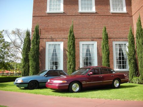 1992 Ford Crown Victoria - Two sedans are parked on a lawn in front of a brick building with large windows.