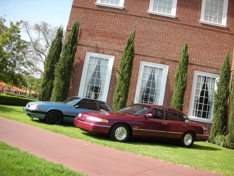 1992 Ford Crown Victoria - Two Ford sedans, a maroon Crown Victoria and a blue Mustang, are parked on a lawn in front of a brick building.
