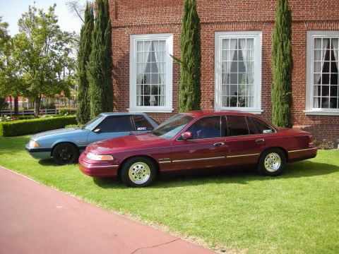 1992 Ford Crown Victoria - Two Ford vehicles are parked on a lawn in front of a brick building with large windows.