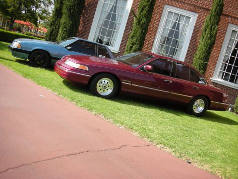 1992 Ford Crown Victoria - A maroon 1992 Ford Crown Victoria is parked on a grassy lawn next to a blue coupe in front of a brick building.