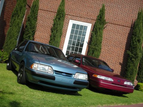 1992 Ford Crown Victoria - Two Ford sedans, a Crown Victoria and a Mustang, are parked on a lawn in front of a brick building.