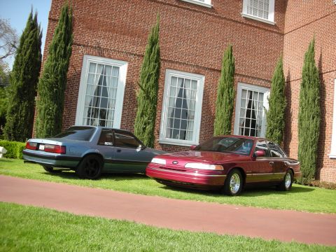 1992 Ford Crown Victoria - Two cars, a red 1992 Ford Crown Victoria and a blue coupe, are parked on the lawn in front of a large brick building.