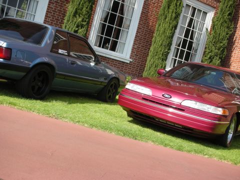 1992 Ford Crown Victoria - Two Ford vehicles, a Crown Victoria and a Mustang, parked on the lawn in front of a brick building.