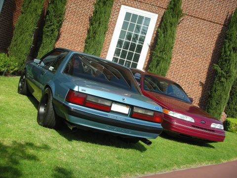 1992 Ford Crown Victoria - Two cars are parked on a grassy area in front of a brick building with a large window.