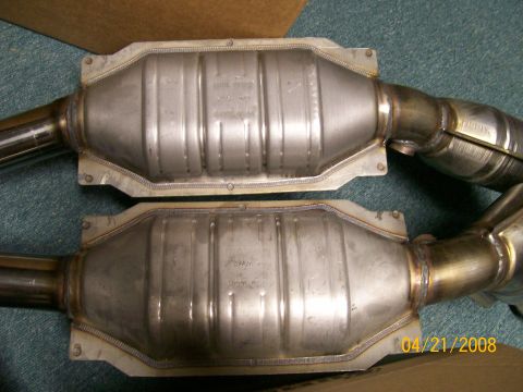1995 Mercury Grand Marquis - A matched pair of stainless dual catalytic converters with welded inlet and outlet pipes, laid side by side on green carpet next to a cardb…