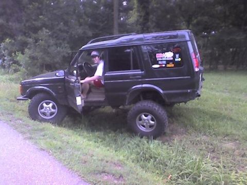 1995 Ford Bronco - A black Land Rover Discovery is parked on a grassy incline with its driver's door open and a person seated inside.