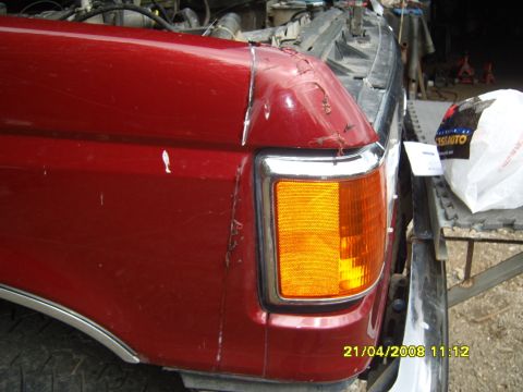 1989 Ford Bronco - Close-up of a dark red front fender corner with a chrome-trimmed amber corner marker lamp, showing cracked, chipped paint and body filler d…