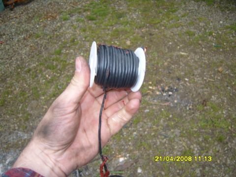 1989 Ford Bronco - A grease-stained hand holds a small white plastic spool of black primary wire outdoors over gravel and patchy grass, with the loose lead tr…