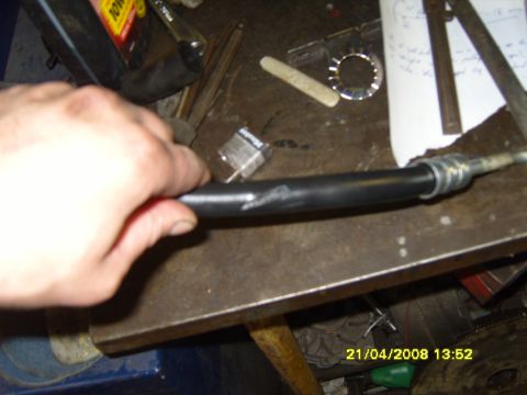 1989 Ford Bronco - A hand grips a black hose with metal fittings, surrounded by tools and papers on a workbench.