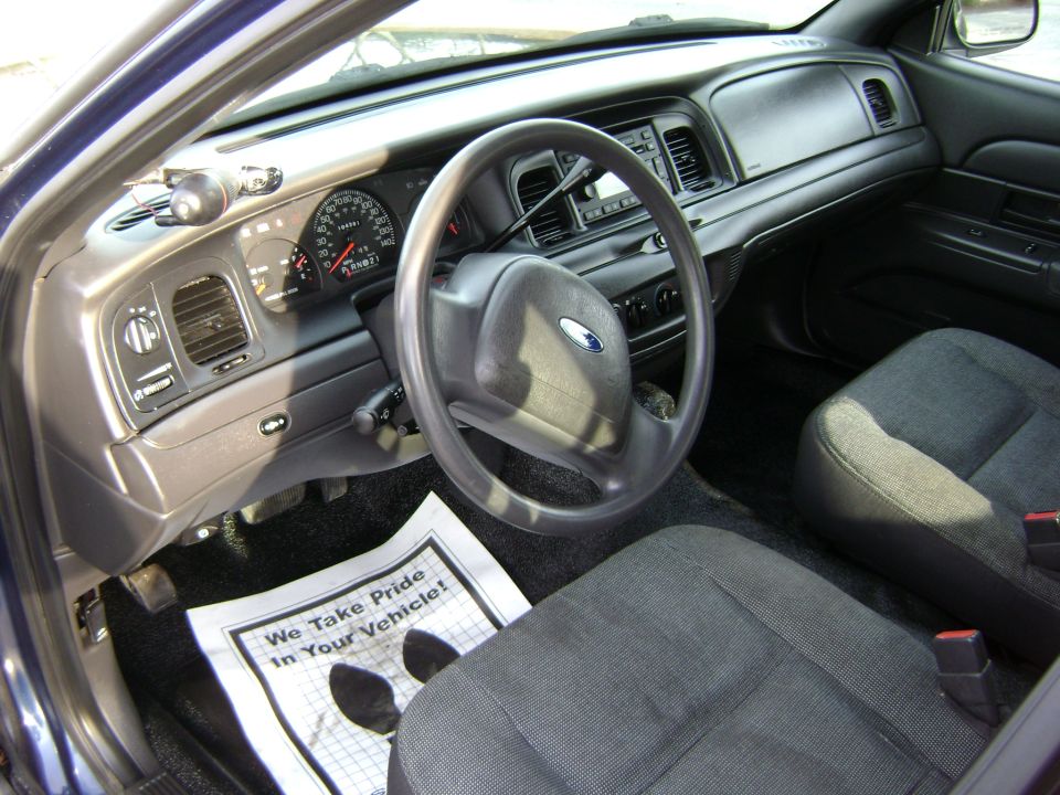 Interior of a 2001 Ford Crown Victoria showing the dashboard, steering wheel, and front seats. A paper on the floor reads 'We Take Pride In Your Vehicle!'