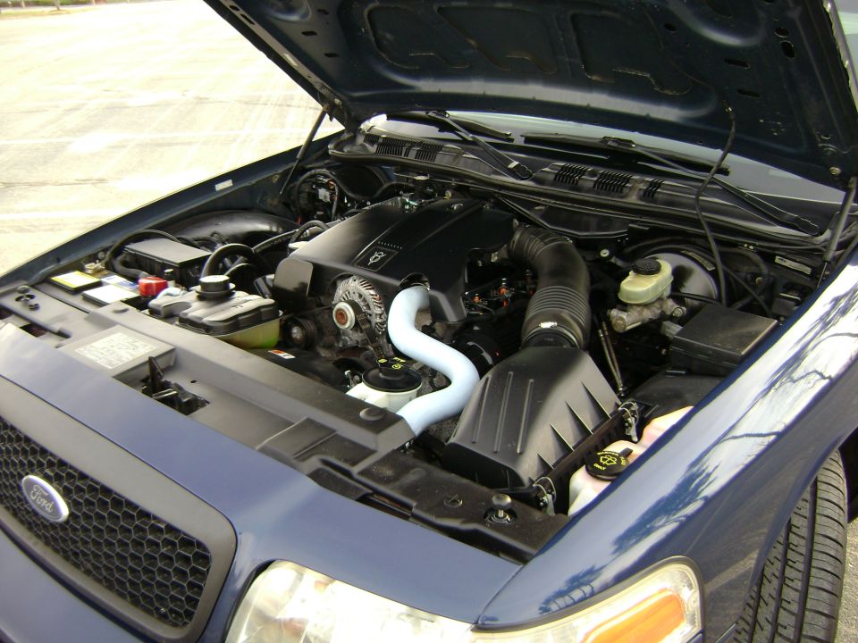 2001 Ford Crown Victoria engine bay with hood open. Black engine, white coolant hose, and blue exterior paint visible