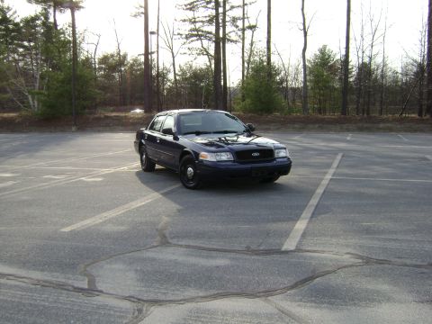 2001 Ford Crown Victoria - A dark blue 2001 Ford Crown Victoria is parked in a lot with trees in the background.