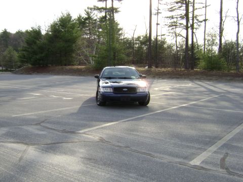 2001 Ford Crown Victoria - A dark blue 2001 Ford Crown Victoria is parked in a paved lot with trees in the background.