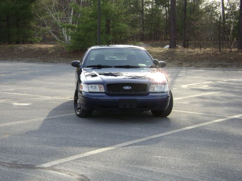 2001 Ford Crown Victoria - A dark blue 2001 Ford Crown Victoria is parked in an empty lot with trees in the background.