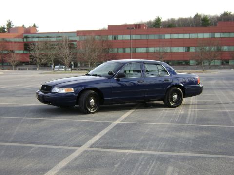 2001 Ford Crown Victoria - A dark blue 2001 Ford Crown Victoria sedan is parked in an asphalt lot with a modern office building in the background.