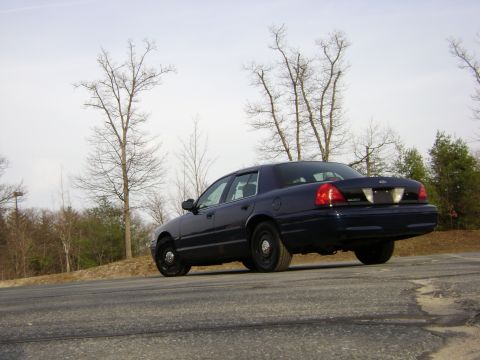 2001 Ford Crown Victoria - A dark blue 2001 Ford Crown Victoria is parked on a road with trees in the background.
