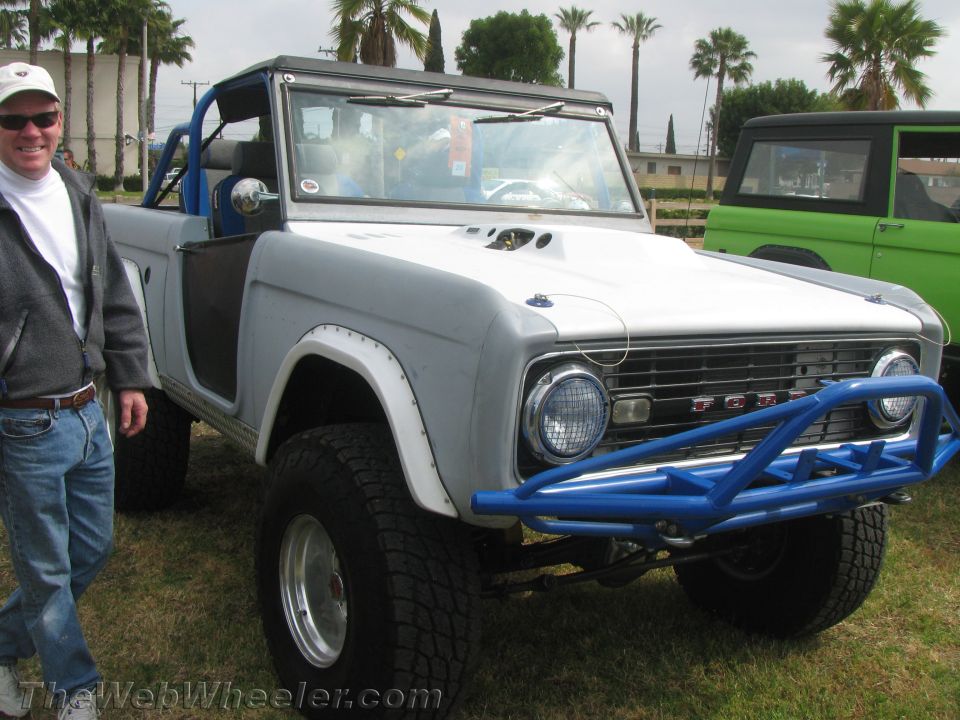 Gray Ford Bronco with blue roll cage and custom bumper, parked next to a green Bronco