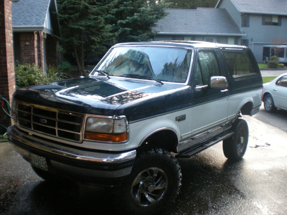 Two-tone dark green and white Ford Bronco with aftermarket wheels and tires, lifted suspension, parked on wet pavement
