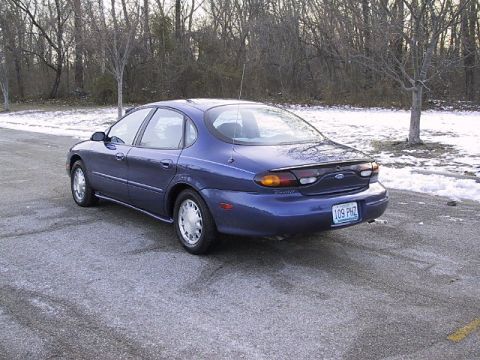 1996 Ford Taurus - A blue 1996 Ford Taurus sedan is seen from the rear quarter angle on a paved road with snow on the ground.