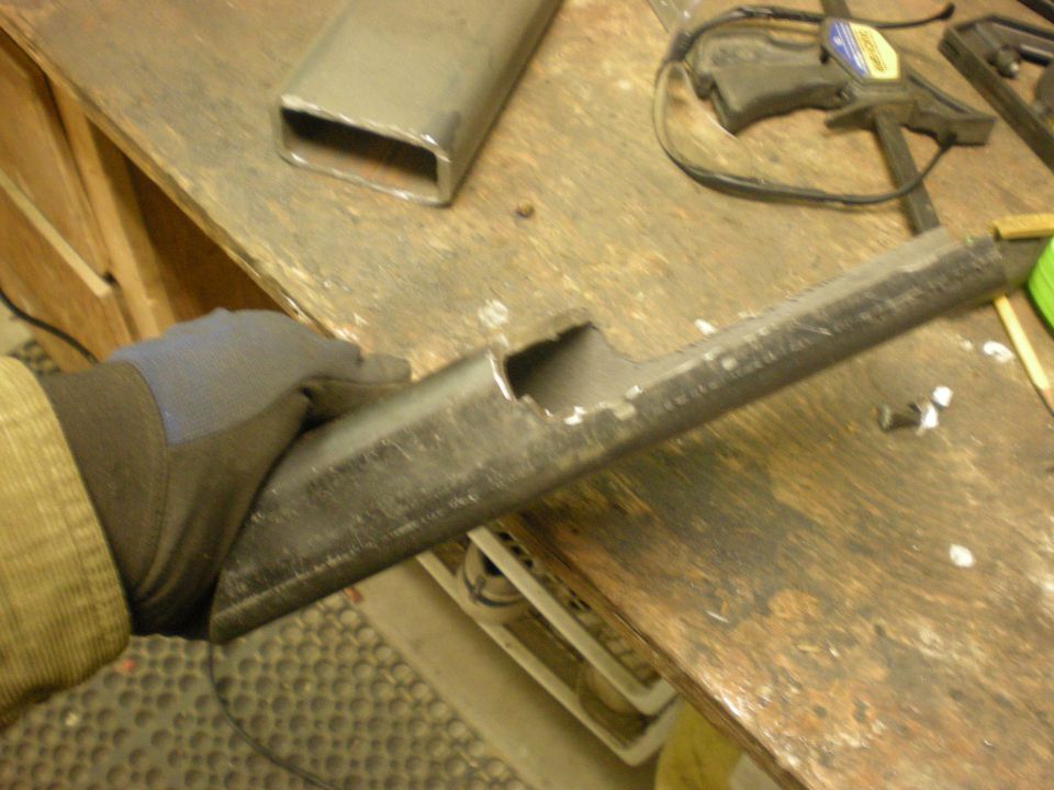 Gloved hand holding a metal tube with a rectangular cut-out, resting on a wooden workbench