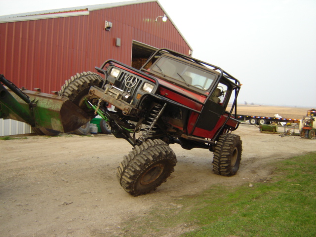 Modified red Jeep Wrangler with large tires and a roll cage, interacting with a green front loader. Farm building in background