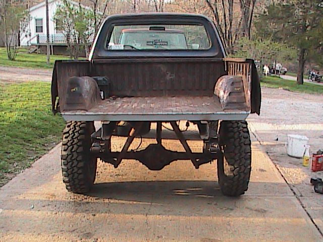 Rear view of a lifted pickup truck with large tires and a worn bed. Another vehicle is visible through the rear window