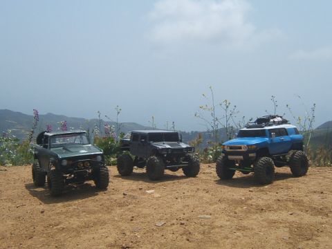 1988 Ford Bronco - Three remote-controlled off-road vehicles are parked on a dirt path with a mountainous landscape behind them.