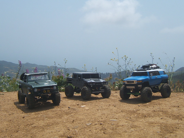 Three RC off-road vehicles on a dirt path with mountains in the background. Green, black, and blue vehicles