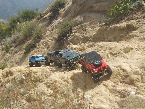 1988 Ford Bronco - Three remote-controlled off-road vehicles navigate a steep, rocky trail under a bright sky.